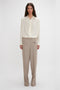 A person stands against a plain background, wearing the Victoria Beckham Asymmetric Ruffle Blouse In Ivory with voluminous sleeves and a ruffled front, paired with beige trousers featuring a button-front closure.