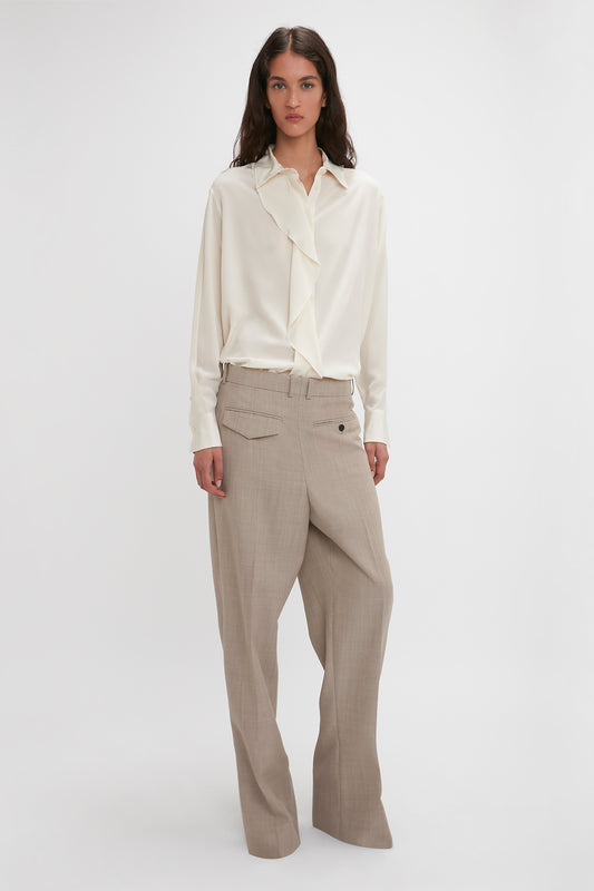 A person wearing a Victoria Beckham Asymmetric Ruffle Blouse In Ivory with voluminous sleeves and loose-fitting beige trousers stands against a plain white background.