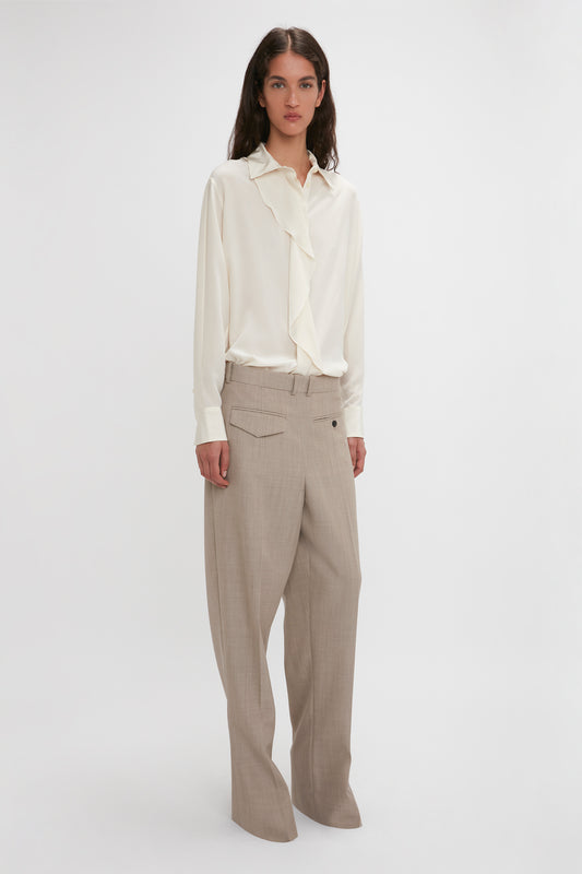 A person with long hair is wearing an Asymmetric Ruffle Blouse In Ivory by Victoria Beckham with voluminous sleeves and beige wide-legged trousers, standing against a plain white background.