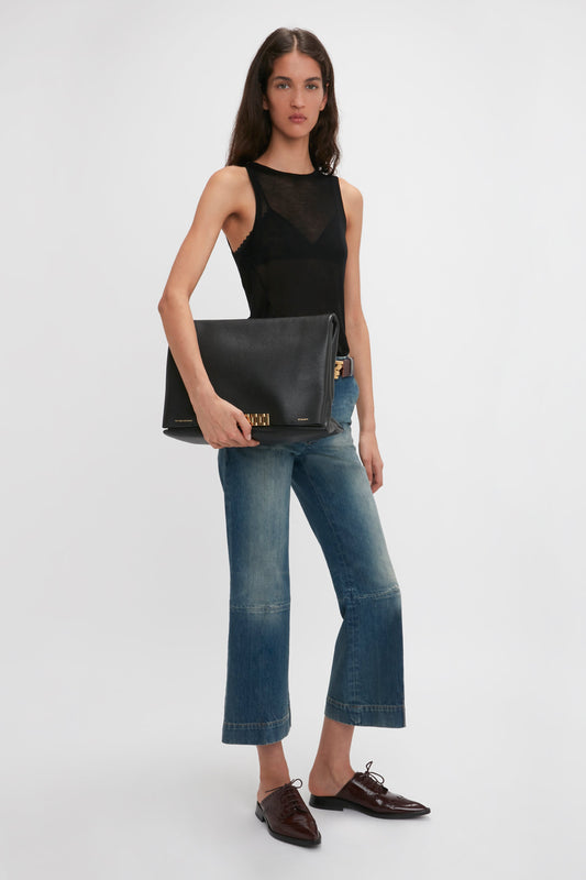 A person with long hair wearing a Victoria Beckham Lightweight Tank Top In Black, blue jeans, and brown shoes holds a large black Givenchy bag against a white background.