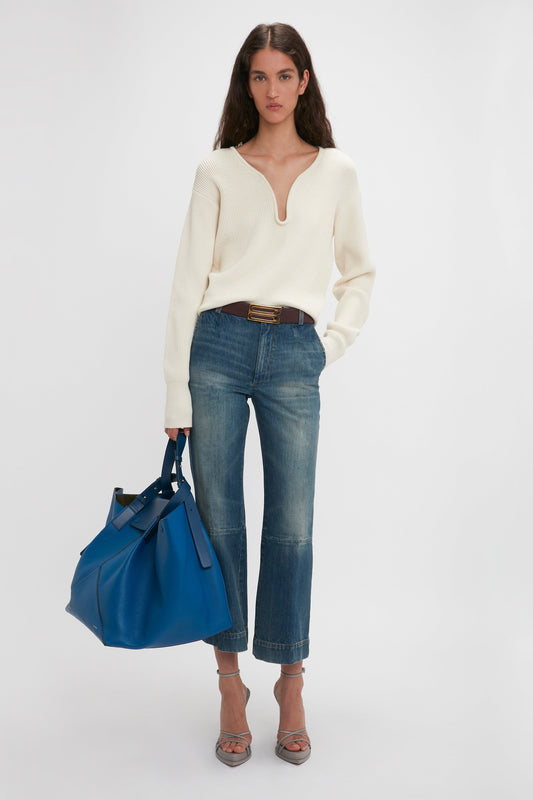 A woman stands against a plain backdrop, wearing a white long-sleeve top, blue jeans, and heeled sandals, holding a Victoria Beckham W11 Medium Tote Bag In Vibrant Blue.