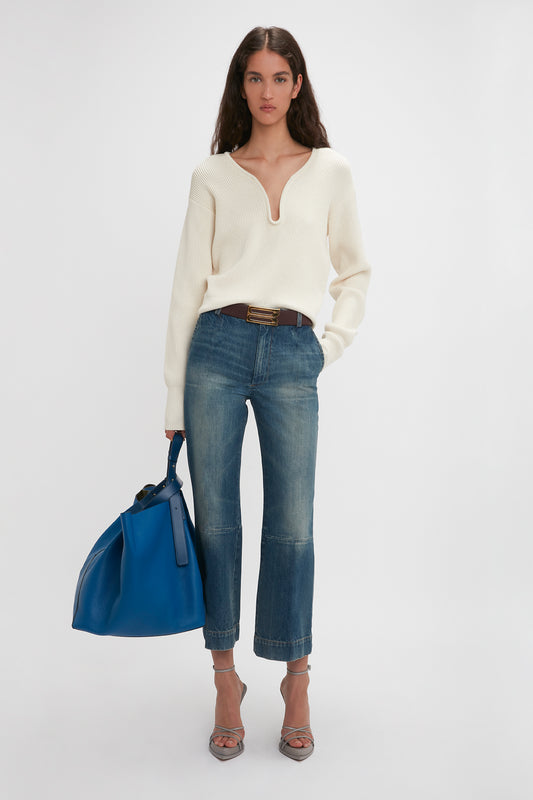 A woman stands against a plain background, wearing a cream sweater, Victoria Beckham Flared Silhouette Blue Jeans, and holding a blue bag, with heels.