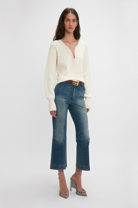 A woman in a white sweater and Victoria Beckham Cropped Kick Jean in Indigrey Wash stands against a white background, accessorized with a belt and heeled sandals.
