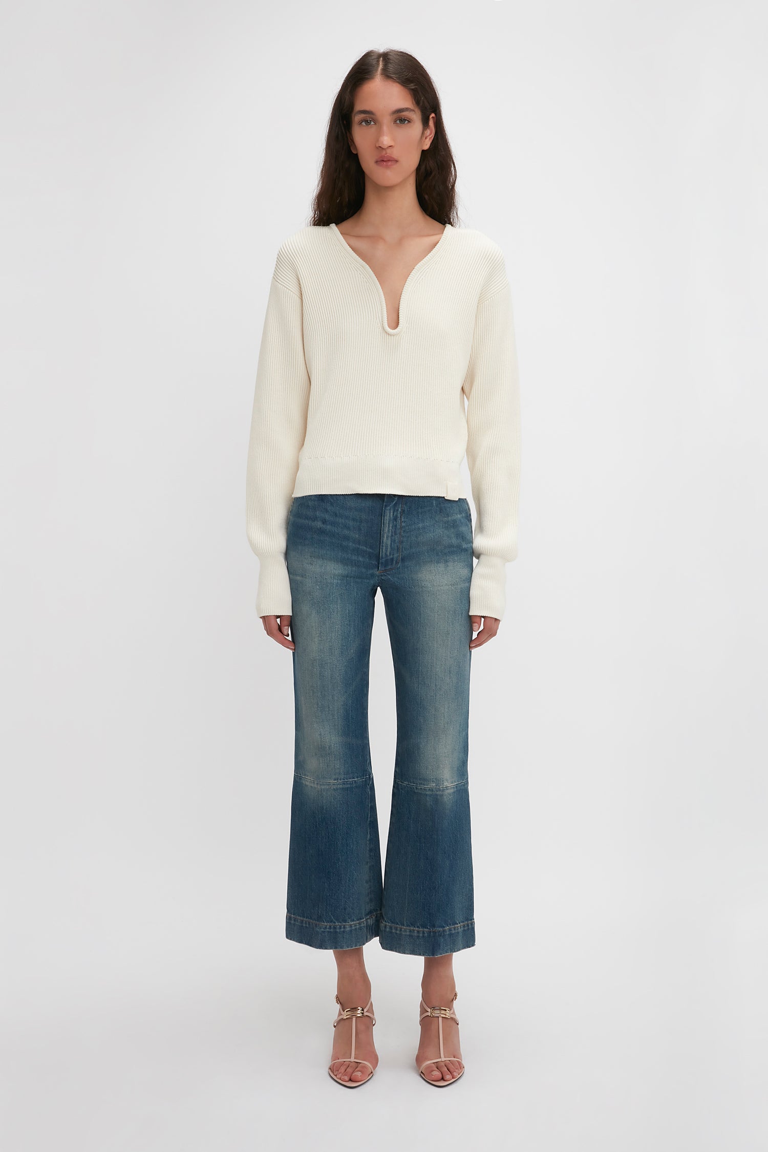 A woman standing against a plain white background wears a cream-colored long-sleeve top, blue wide-leg jeans, and Victoria Beckham Frame Detail Sandal In Nude Leather with adjustable ankle straps.