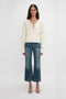 A woman standing against a plain white background wears a cream-colored long-sleeve top, blue wide-leg jeans, and Victoria Beckham Frame Detail Sandal In Nude Leather with adjustable ankle straps.