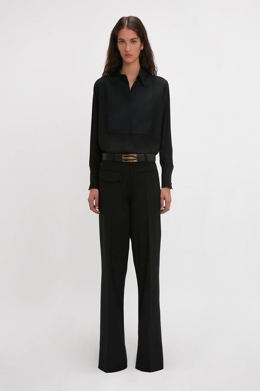 A modern woman with long hair stands facing forward, wearing a black long-sleeve shirt and Victoria Beckham's Reverse Front Trouser In Black, showcasing a contemporary silhouette.