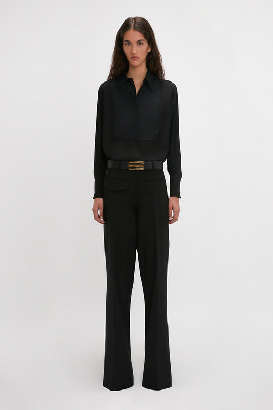 Person stands against a plain background wearing a timeless black **Contrast Bib Shirt In Black** by **Victoria Beckham** and high-waisted pants with a belt.