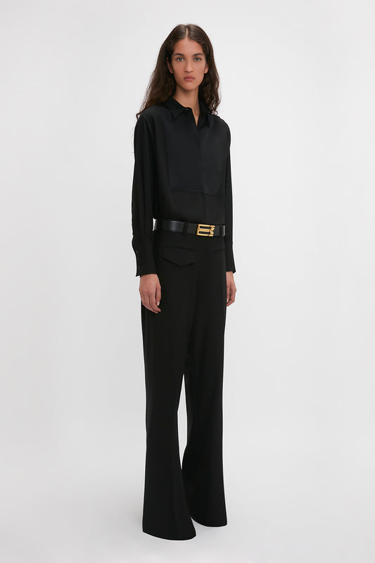 A person with long, dark hair stands against a plain background, wearing a timeless Victoria Beckham Contrast Bib Shirt In Black with a collar, black high-waisted pants, and a black belt with a gold buckle, exuding modern femininity.