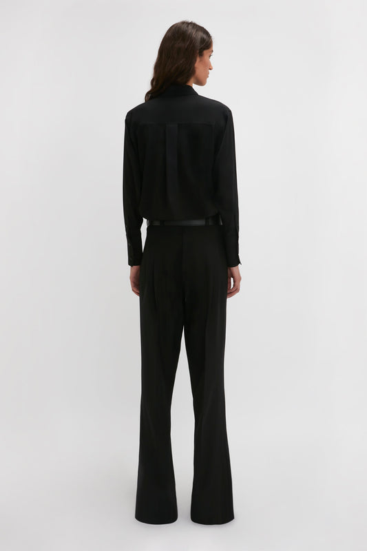 A contemporary silhouette of a modern woman in a black long-sleeve shirt and Victoria Beckham’s Reverse Front Trouser In Black, facing away against a plain white background.