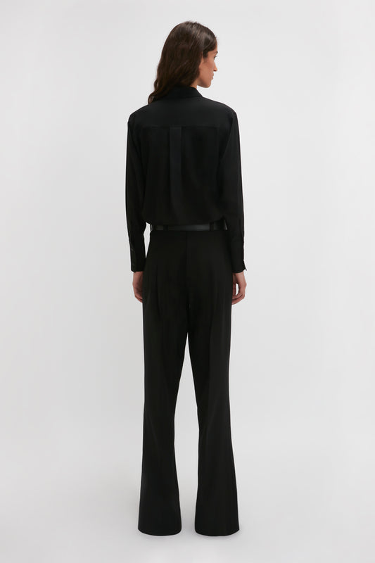 A person with long hair, dressed in a timeless Victoria Beckham Contrast Bib Shirt In Black and black pants, is standing with their back facing the camera against a plain white background.