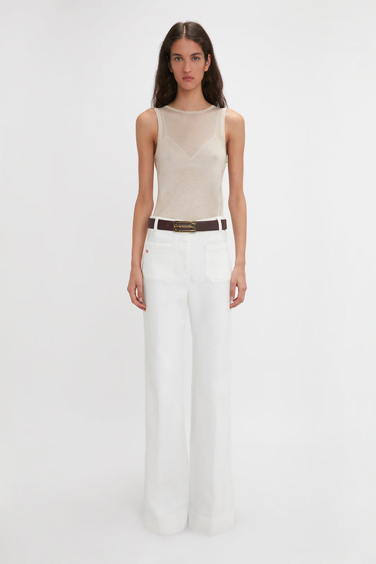 A person stands against a plain background wearing a Victoria Beckham Lightweight Tank Top in Birch, white high-waisted wide-leg pants, and a dark brown belt.