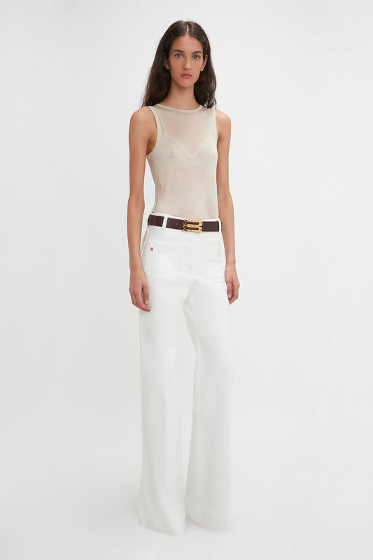 A person stands against a plain white background wearing a Victoria Beckham Lightweight Tank Top In Birch, wide-leg white pants, and a brown belt with a gold buckle. They have long, dark hair and a neutral expression.