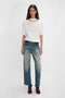 The woman stands against a plain background, wearing a relaxed cut Bonded Jersey Tee In White by Victoria Beckham with dropped shoulders, faded blue jeans, a black belt, and dark brown ankle boots.