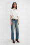 A person standing against a plain background wears a white Victoria Beckham Bonded Jersey Tee In White with a cutout design and relaxed cut, high-waisted blue jeans, a black belt with gold buckle, and dark red high-heeled shoes.