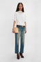 A person stands against a plain background, wearing a white Bonded Jersey Tee In White by Victoria Beckham with dropped shoulders, wide-leg faded jeans, and burgundy shoes with heels, holding a beige clutch bag.