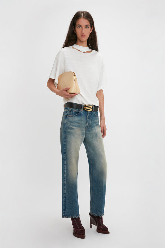 Woman with long hair wearing a white t-shirt, faded blue jeans, burgundy high heels, and a black belt. She holds the Victoria Clutch Bag In Sesame Leather by Victoria Beckham and stands against a plain white background.