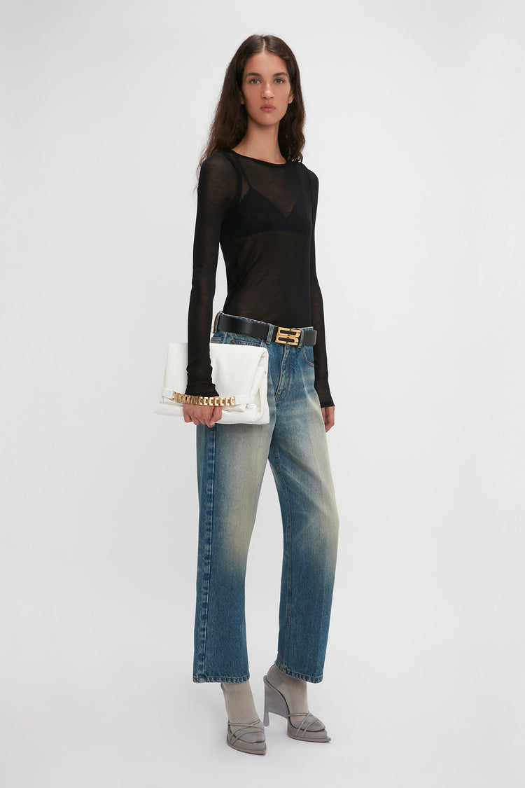 A person with long hair wears a black Crew Neck Long Sleeve Top In Black from Victoria Beckham, blue jeans, and grey heels, holding a white clutch bag against a plain background.