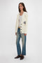 A person with long hair is wearing a Victoria Beckham Relaxed Fit Cardigan In Natural, along with a white top, blue jeans, and brown shoes, standing against a plain white background.