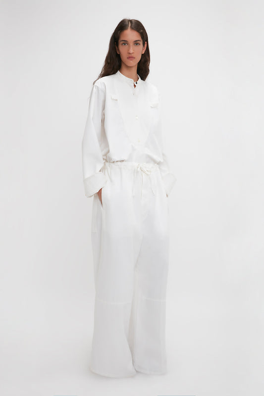 A woman in stylish Victoria Beckham white cotton-canvas drawstring pyjama trousers stands against a plain background, looking directly at the camera.