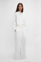 A woman in stylish Victoria Beckham white cotton-canvas drawstring pyjama trousers stands against a plain background, looking directly at the camera.