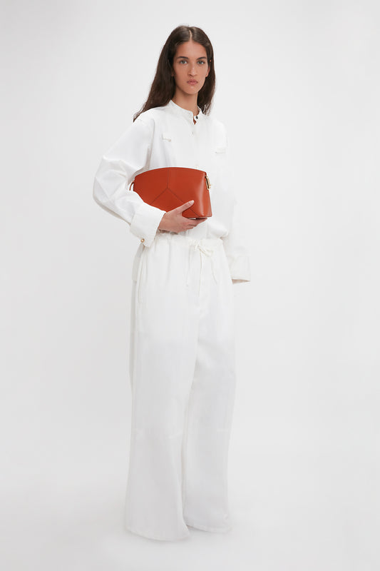 Person in a white outfit holding a Victoria Beckham Victoria Clutch Bag In Tan Leather against a plain white background.