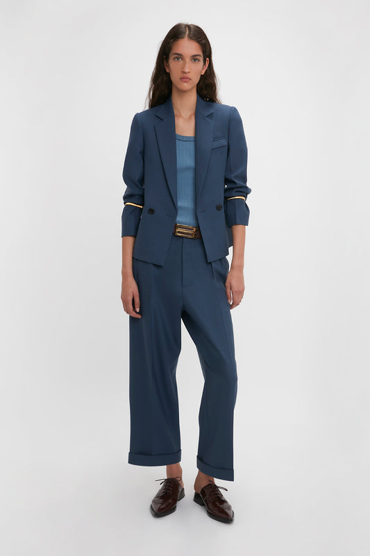 A woman in a Victoria Beckham Heritage Blue shrunken double-breasted jacket and brown shoes stands against a white background, looking directly at the camera.