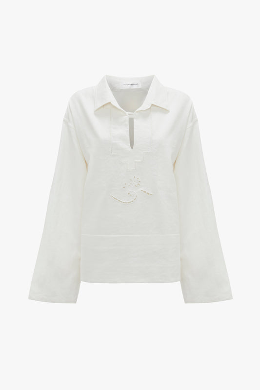 The Victoria Beckham Oversized Embroidered Tunic In Antique White is a vintage-inspired white long-sleeved blouse with a collar and a small front cut-out. Decorative stitching is present on the front, adding to its charm. Crafted from a linen-cotton blend, this relaxed-fit tunic offers both style and comfort.