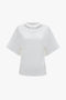 A plain white Bonded Jersey Tee In White by Victoria Beckham with a round neckline, dropped shoulders, and a slightly loose fit on a plain backdrop.