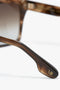 Close-up of the arm of a pair of Victoria Beckham Soft Square Frame Sunglasses In Striped Khaki Havana with the letters "IB" engraved in gold. The frame is brown with a tortoiseshell pattern and gradient lenses, showcasing a stylish and sophisticated design.