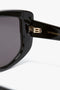 Close-up of Acetate Visor Sunglasses In Black with dark, tonal lenses. The oversized frames feature gold hinges on the arms, each inscribed with "Victoria Beckham.