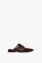 A brown, slip-on leather shoe with a low heel and decorative brogue detailing, crafted from luxurious calfskin leather. This stylish footwear features a closed toe and an open back, perfect for adding elegance to any outfit. The Victoria Beckham Flat Lace Up Mules In Bordeaux Leather embody sophistication and style in every detail.