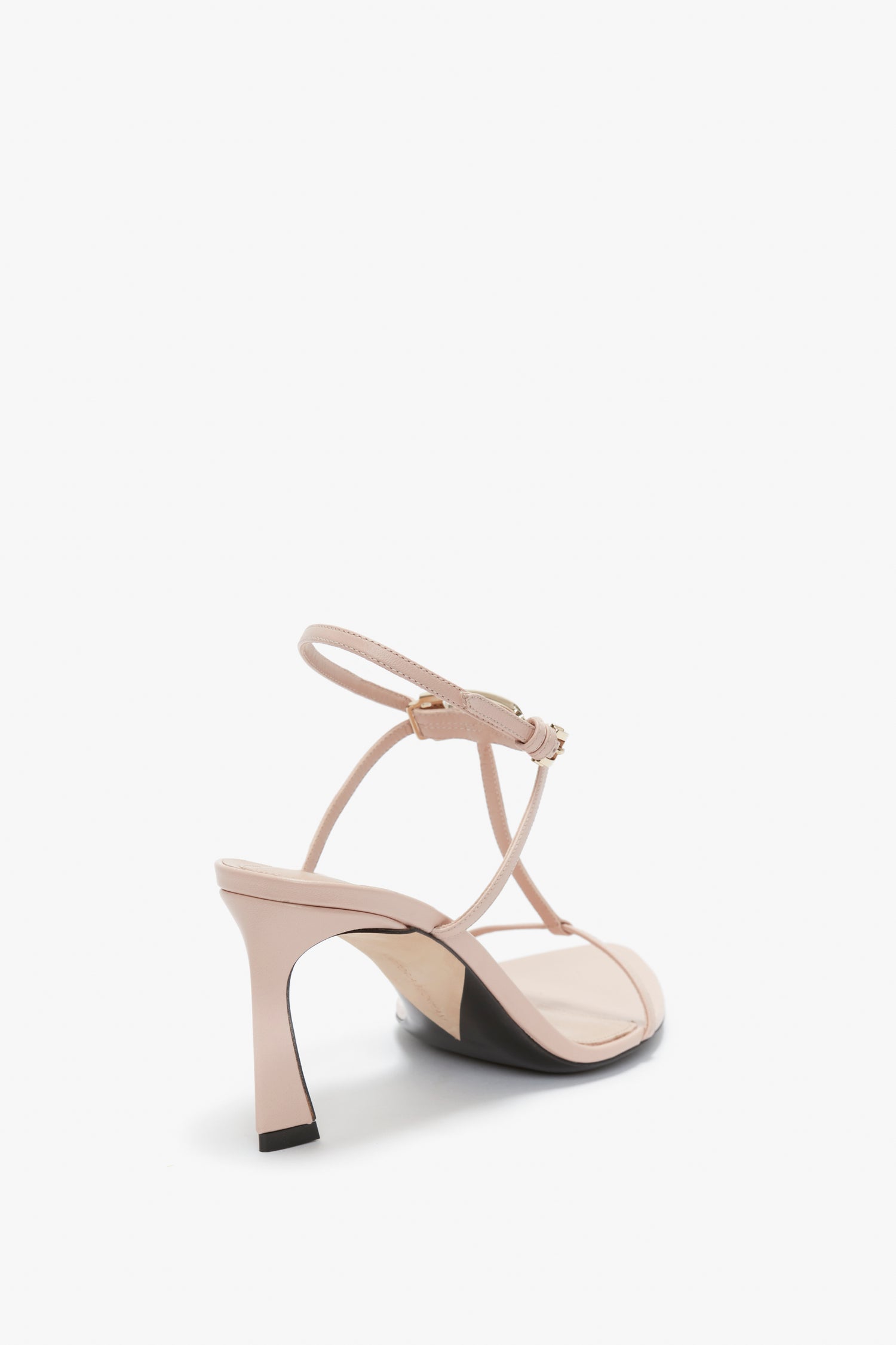 A Victoria Beckham Frame Detail Sandal In Nude Leather with a V-shaped sculptural heel is pictured against a plain white background. The light beige high-heeled sandal features a thin strap, an adjustable ankle strap, and a gold buckle.