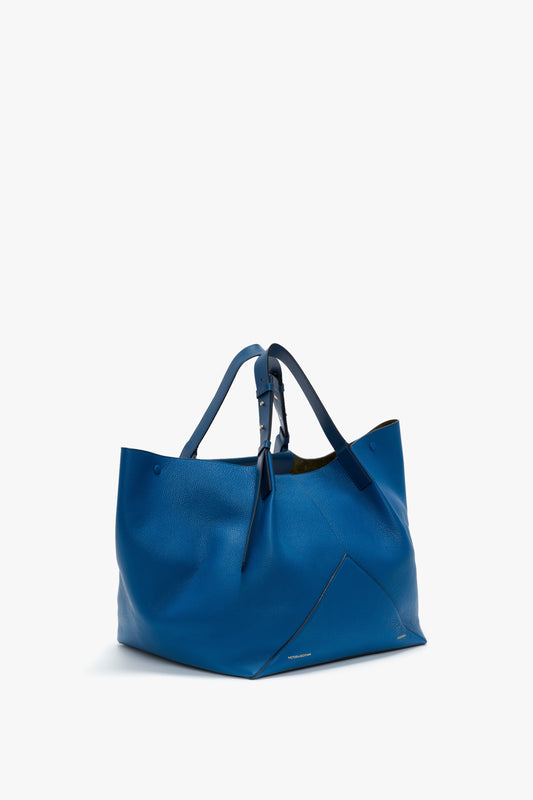 A W11 Medium Tote Bag In Vibrant Blue made from grained leather with two adjustable shoulder straps and a tie closure, placed on a white background, by Victoria Beckham.