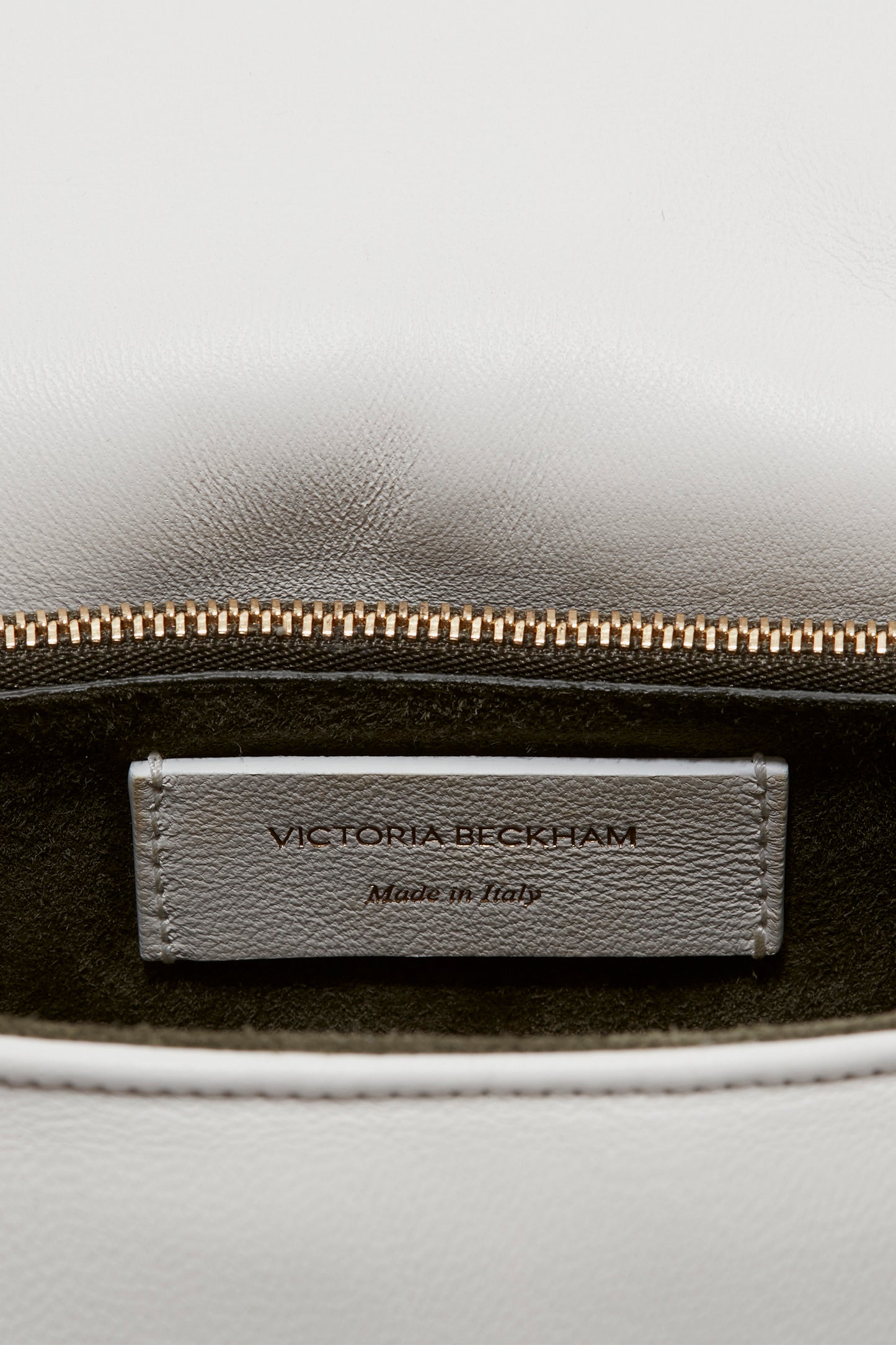 Close-up of a Victoria Beckham Puffy Chain Pouch Bag With Strap In White Leather's interior label with the text "VICTORIA BECKHAM Made in Italy," shown below a partially open brass-colored zipper. The fabric is white and leather-like.