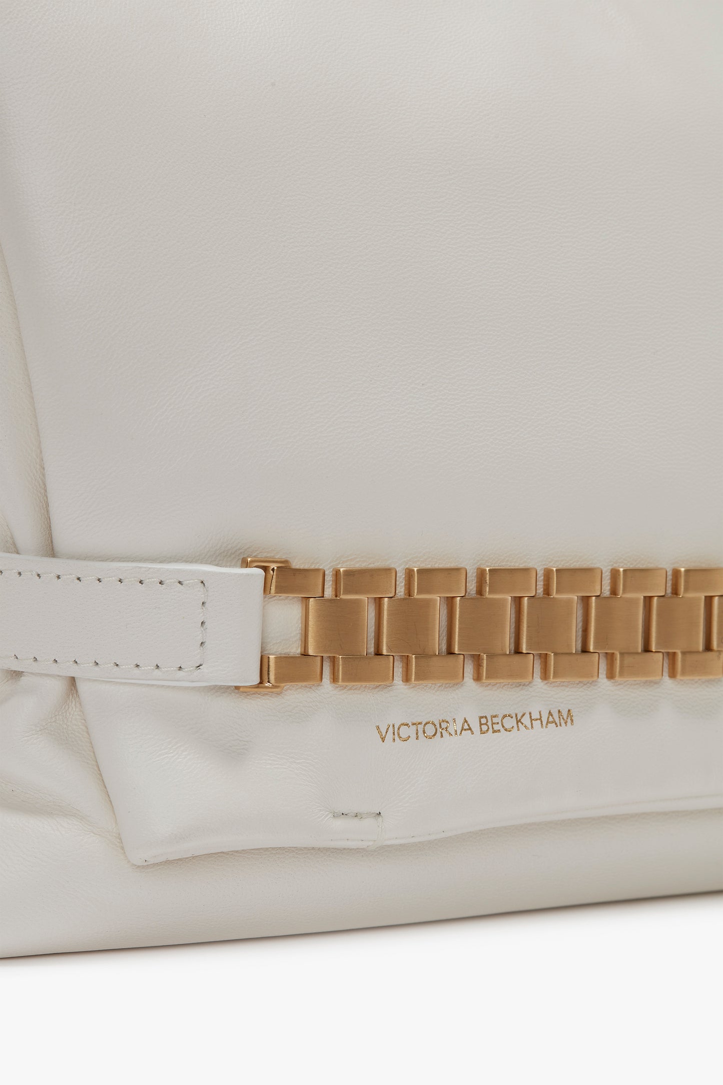 Close-up of a VB handbag, featuring a white Puffy Chain Pouch Bag With Strap In White Leather with gold chain detail and "Victoria Beckham" embossed in gold on the lower corner.