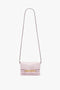 A small, light pink Mini Chain Pouch Bag With Long Strap In Orchid Leather by Victoria Beckham with a decorative gold clasp on the front and a long, thin strap.