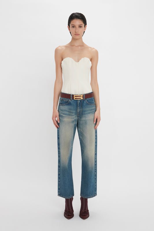 A woman modeling a Victoria Beckham Corset Top In Antique White and blue jeans, accessorized with a brown belt, standing against a white background.