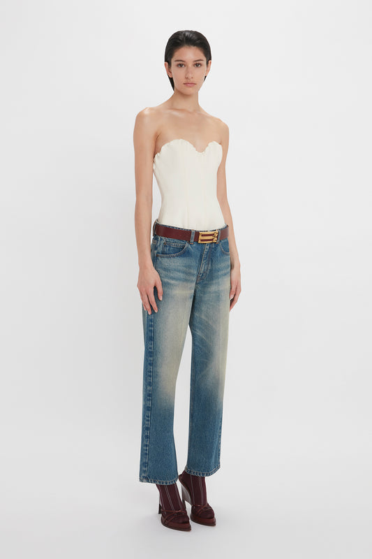 A woman in a Victoria Beckham corset top in antique white and blue jeans, accessorized with a brown belt, standing against a plain background.