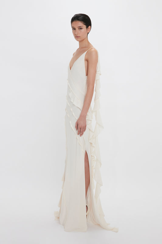 A woman stands against a white background, wearing an elegant off-white gown with asymmetrical ruffled layers and a crepe back satin high slit by Victoria Beckham's Exclusive Asymmetric Bias Frill Dress In Ivory.