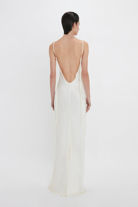 A woman stands facing away from the camera, wearing an elegant Victoria Beckham Exclusive Lace Detail Floor-Length Cami Dress in Ivory with a deep back and thin straps.