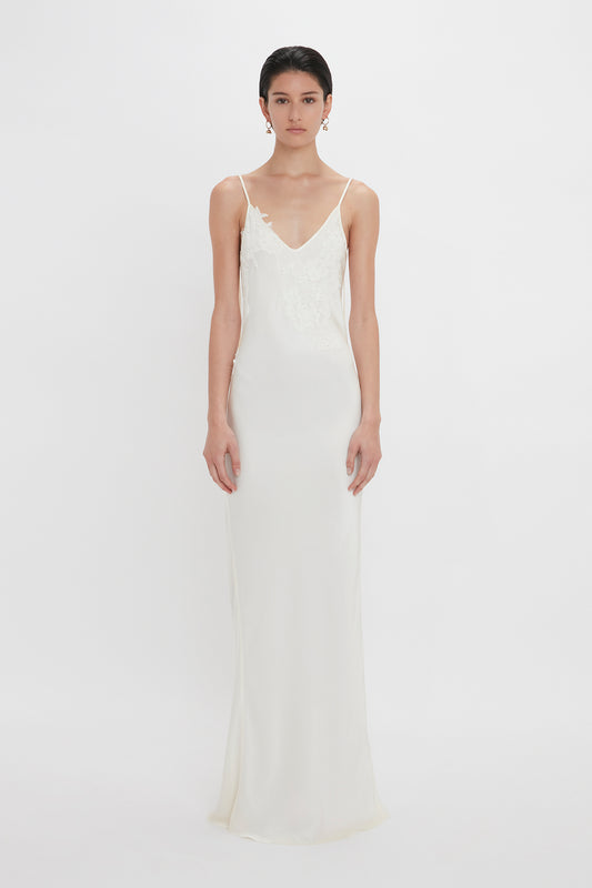 A woman stands against a white background wearing an elegant, sleeveless white wedding dress with lace appliqué detail on the bodice by Victoria Beckham.