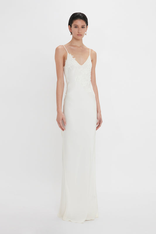 A woman stands against a white background, wearing a slim-fit, white sleeveless wedding gown with lace appliqué detail. This is the Exclusive Lace Detail Floor-Length Cami Dress in Ivory by Victoria Beckham.