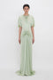 A woman models a pale green, elegant, Exclusive Floor-Length Gathered Dress In Jade by Victoria Beckham with a gathered twist at the waist and short sleeves, standing against a white backdrop.