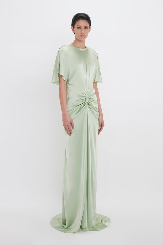 A woman standing in a studio wears an elegant Victoria Beckham Exclusive Floor-Length Gathered Dress In Jade with short sleeves and a gathered twist at the waist.