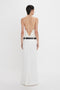 A person is seen from the back wearing an ivory dress with a low-cut back and black belt, standing against a plain white background. The dress features an Exclusive Tailored Floor-Length Pleated Skirt In Ivory by Victoria Beckham, adding elegance to the simple setting.