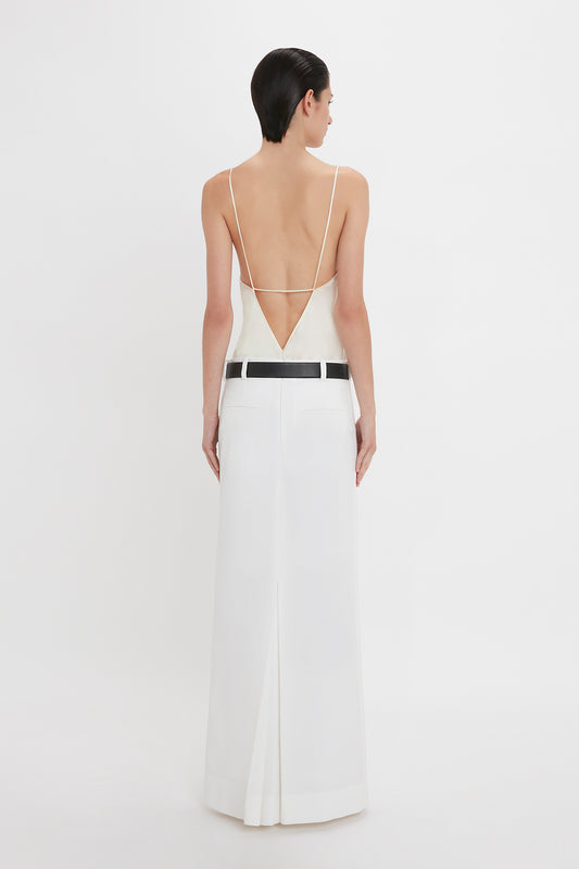 A person stands facing away, wearing the Victoria Beckham Exclusive Cami Top In Ivory with thin straps and an open back, as well as white pants with a wide black belt.