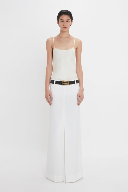 A person stands against a white background, wearing an Exclusive Cami Top In Ivory by Victoria Beckham and a long white skirt, cinched with a black belt featuring a gold buckle. Their hands rest by their sides, adding to the elegant simplicity of the look.