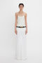 A person stands against a white background, wearing an Exclusive Cami Top In Ivory by Victoria Beckham and a long white skirt, cinched with a black belt featuring a gold buckle. Their hands rest by their sides, adding to the elegant simplicity of the look.