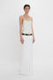 A person with short hair wearing an ivory sleeveless top and an Exclusive Tailored Floor-Length Pleated Skirt In Ivory by Victoria Beckham, accessorized with a black belt, stands against a plain white background.