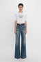 A person standing against a white background, wearing a white T-shirt with the text "DAVID'S WIFE" and blue Alina High Waisted Jean In Indigrey Wash by Victoria Beckham.